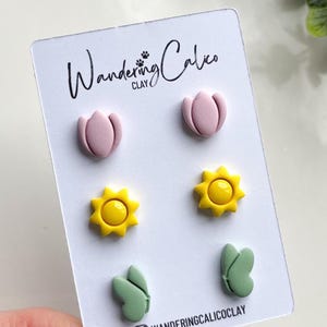 Puede incluir: Un conjunto de seis pendientes de arcilla polim&eacute;rica con una flor rosa, un sol amarillo y una hoja verde. Los pendientes se muestran en una tarjeta blanca con el texto "Wandering Calico Clay".