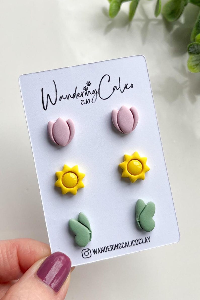Puede incluir: Un conjunto de seis pendientes de arcilla polim&eacute;rica con una flor rosa, un sol amarillo y una hoja verde. Los pendientes se muestran en una tarjeta blanca con el texto "Wandering Calico Clay".