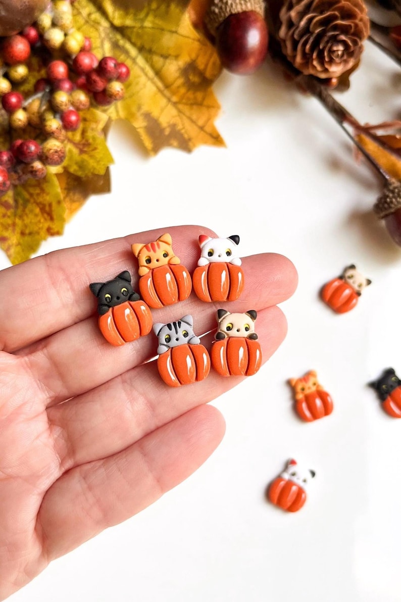 May include: A collection of miniature cat figurines nestled in orange pumpkin shapes. The cats have various colors and patterns, including black, gray, and orange tabby. These fall-themed decorations are perfect for seasonal crafts or home decor.