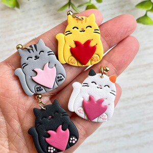 May include: Four pairs of cat-shaped earrings with different colored fur and a pink heart. The cats are holding the hearts in their paws.
