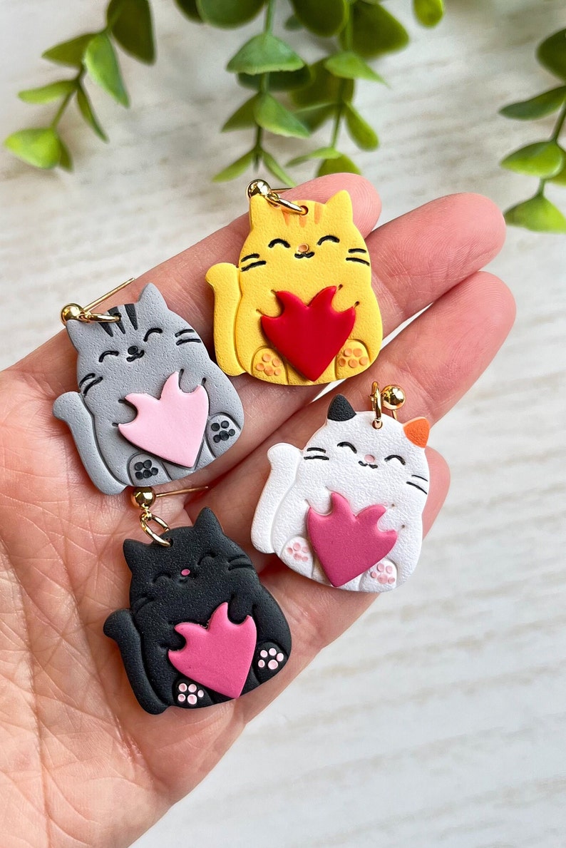 May include: Four pairs of cat-shaped earrings with different colored fur and a pink heart. The cats are holding the hearts in their paws.