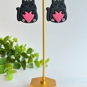 May include: A pair of black cat earrings with pink hearts. The earrings are on a gold stand.