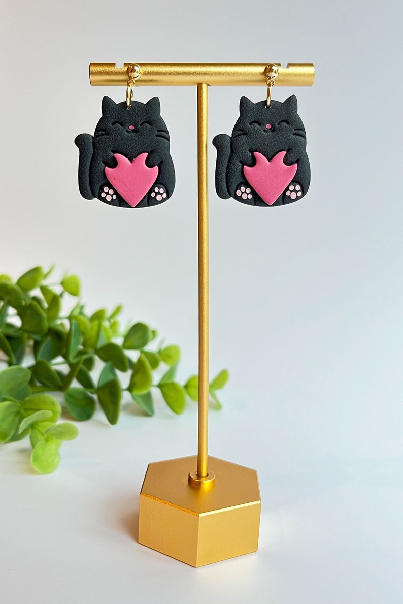 May include: A pair of black cat earrings with pink hearts. The earrings are on a gold stand.