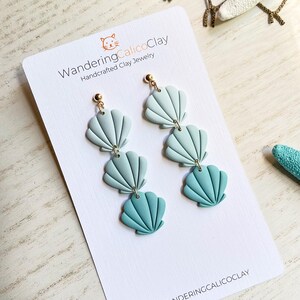 May include: A pair of dangle earrings featuring three layered seashells in shades of blue and green. The earrings are made of polymer clay and have gold-toned hooks. The earrings are displayed on a white card with the text "WanderingCalicoClay Handcrafted Clay Jewelry" and the Instagram handle "@wanderingcalicoclay".