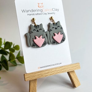 May include: Gray cat holding a pink heart clay earrings with gold hooks. The earrings are displayed on a white card with the text "WanderingCalicoClay Handcrafted Clay Jewelry" and the Instagram handle "@wanderingcalicoclay".