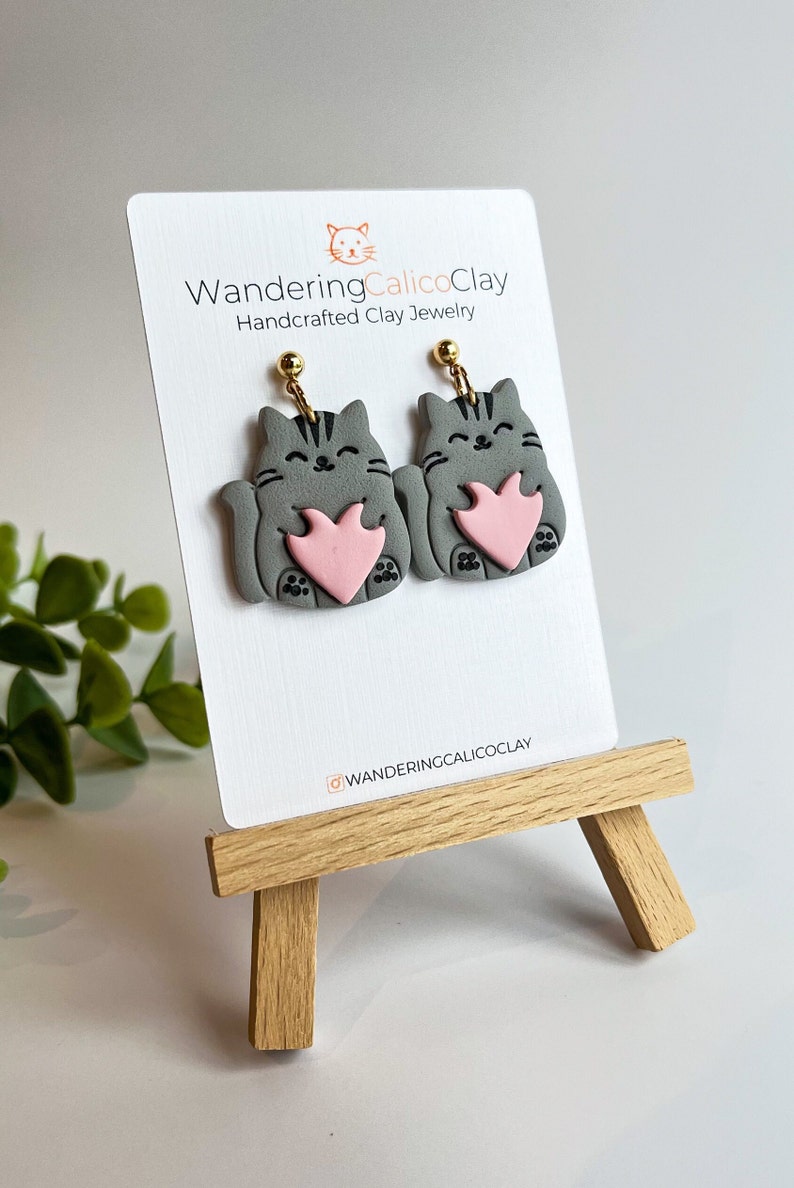 May include: Gray cat holding a pink heart clay earrings with gold hooks. The earrings are displayed on a white card with the text "WanderingCalicoClay Handcrafted Clay Jewelry" and the Instagram handle "@wanderingcalicoclay".