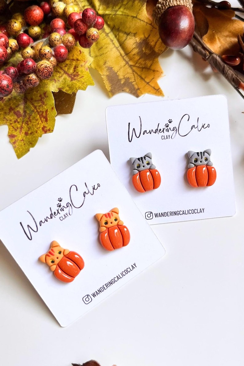 May include: Handmade cat-shaped earrings in orange and gray, nestled in orange pumpkins. The earrings are displayed on white cards with the text "Wandering Calico Clay" and an Instagram handle. Autumn-themed jewelry.
