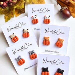 May include: A collection of handmade clay earrings featuring cat figures nestled in orange pumpkins. The earrings are displayed on white cards with the text "Wandering Calico Clay" and the Instagram handle. Autumn-themed jewelry.