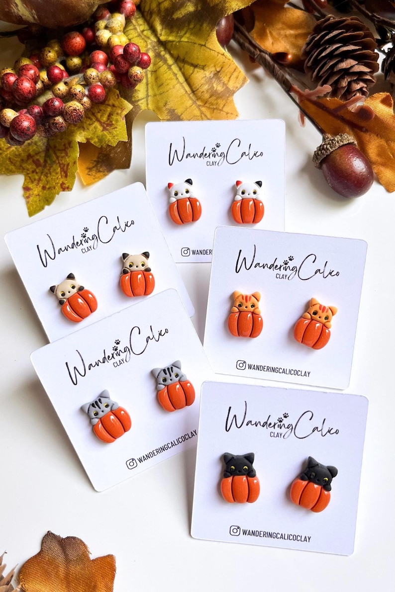 May include: A collection of handmade clay earrings featuring cat figures nestled in orange pumpkins. The earrings are displayed on white cards with the text "Wandering Calico Clay" and the Instagram handle. Autumn-themed jewelry.