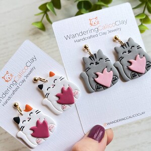 May include: A pair of handmade polymer clay cat earrings. The earrings feature a gray cat holding a pink heart and a white cat holding a burgundy heart. The earrings are on a white card with the text "WanderingCalicoClay Handcrafted Clay Jewelry" and the Instagram handle "@wanderingcalicoclay".