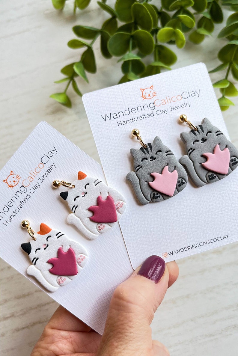 May include: A pair of handmade polymer clay cat earrings. The earrings feature a gray cat holding a pink heart and a white cat holding a burgundy heart. The earrings are on a white card with the text "WanderingCalicoClay Handcrafted Clay Jewelry" and the Instagram handle "@wanderingcalicoclay".