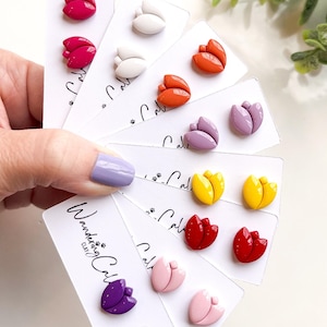 Puede incluir: Un conjunto de 10 pares de pendientes de botón en forma de tulipán en varios colores, incluyendo morado, rosa, rojo, naranja, amarillo y blanco. Los pendientes se muestran en tarjetas blancas con el texto "Wandering Clay Co."