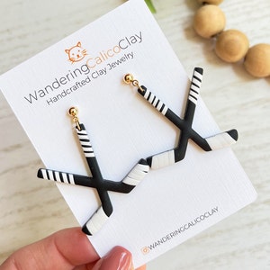 K&ouml;nnte beinhalten: Ein Paar schwarze und wei&szlig;e Hockey-Schl&auml;ger-Ohrringe mit goldenen Stiften. Die Ohrringe werden auf einer wei&szlig;en Karte mit dem Text "WanderingCalicoClay Handcrafted Clay Jewelry" und dem Instagram-Handle "@wanderingcalicoclay" pr&auml;sentiert.