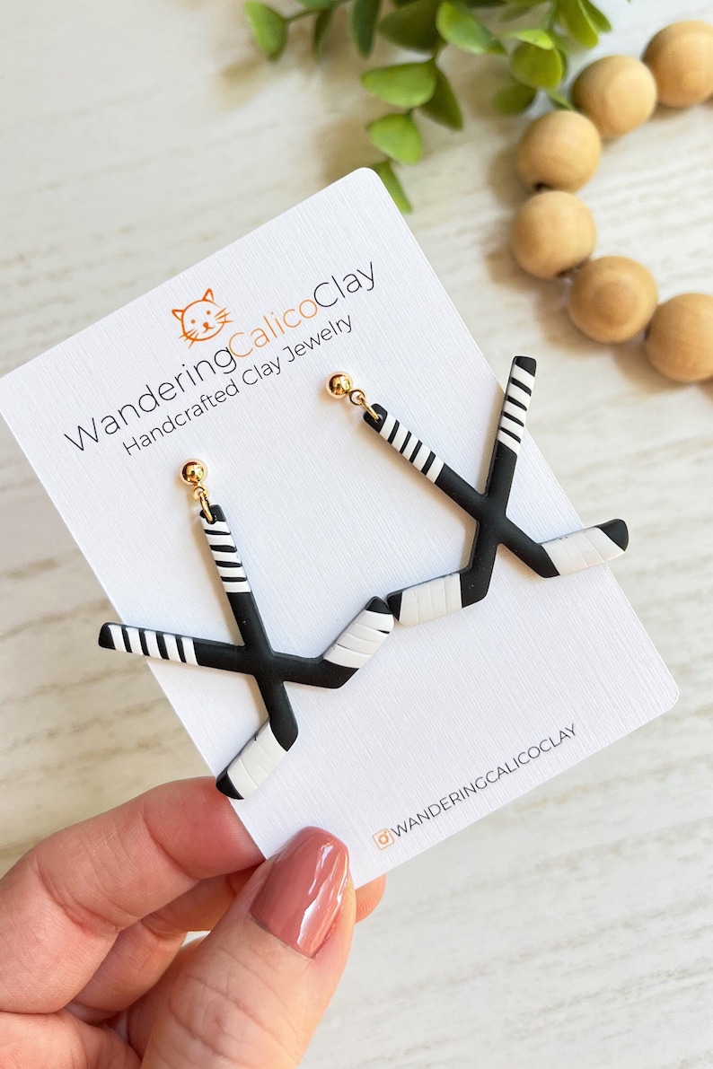 K&ouml;nnte beinhalten: Ein Paar schwarze und wei&szlig;e Hockey-Schl&auml;ger-Ohrringe mit goldenen Stiften. Die Ohrringe werden auf einer wei&szlig;en Karte mit dem Text "WanderingCalicoClay Handcrafted Clay Jewelry" und dem Instagram-Handle "@wanderingcalicoclay" pr&auml;sentiert.