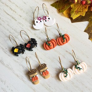 May include: A collection of Halloween-themed earrings. The earrings feature designs such as white ghosts with purple flowers, black spiders with flowers, orange pumpkins, coffee cups with pumpkins, and white pumpkins with green vines. All earrings have gold hoops.
