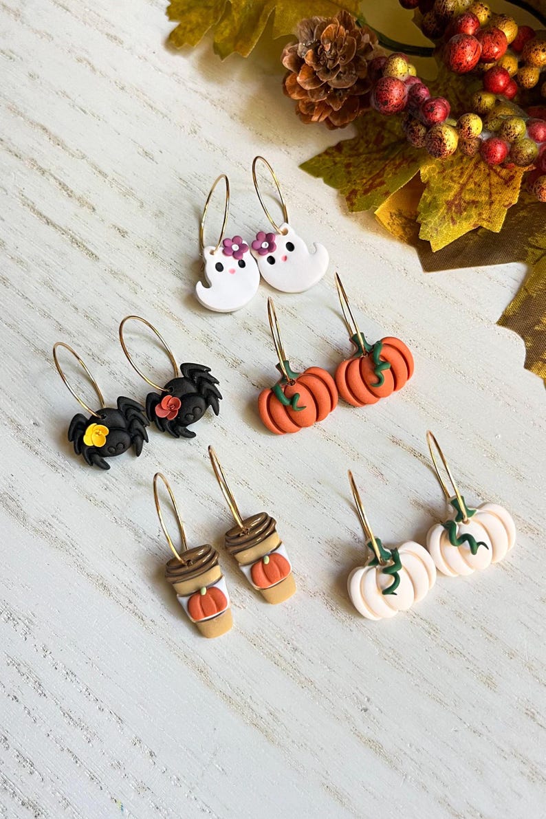 May include: A collection of Halloween-themed earrings. The earrings feature designs such as white ghosts with purple flowers, black spiders with flowers, orange pumpkins, coffee cups with pumpkins, and white pumpkins with green vines. All earrings have gold hoops.