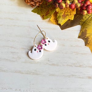 May include: A pair of white ghost-shaped earrings with gold hoops. Each ghost has a small pink flower and black dot eyes. The earrings are set against a white wooden background with fall foliage.