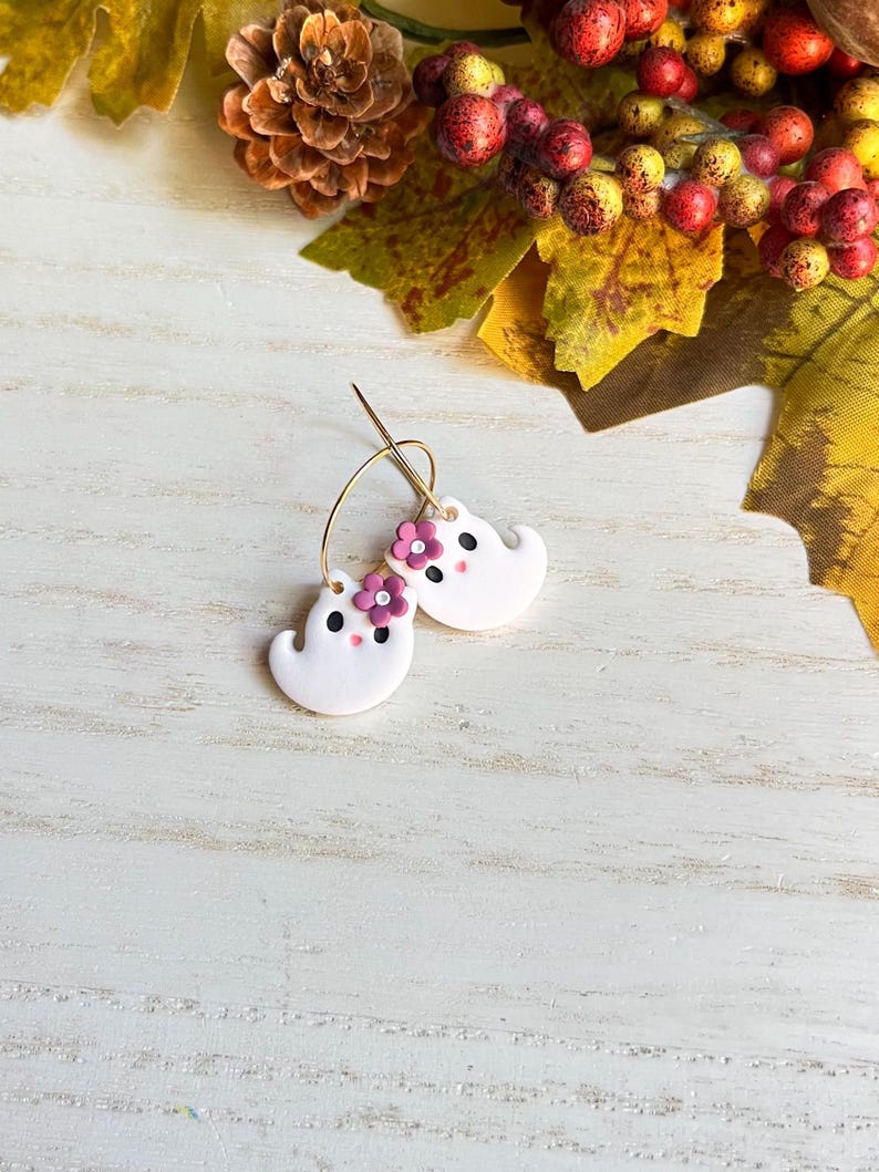 May include: A pair of white ghost-shaped earrings with gold hoops. Each ghost has a small pink flower and black dot eyes. The earrings are set against a white wooden background with fall foliage.