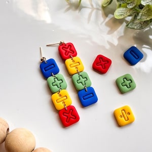 Puede incluir: Un par de pendientes colgantes con una cadena que conecta cuatro cuentas cuadradas de colores. Cada cuenta tiene un símbolo matemático diferente: más, menos, dividir y multiplicar. Las cuentas son rojas, amarillas, verdes y azules.
