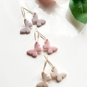 May include: Three pairs of butterfly earrings with gold hoops. The butterflies are made of clay and come in three colours: light pink, light beige, and a darker shade of pink.