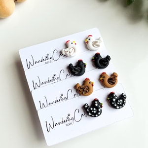 May include: A collection of handmade chicken-shaped earrings in various colors, including white, black, and brown, with red combs and yellow beaks. The earrings are displayed on white cards with the text "Wandering Clay".