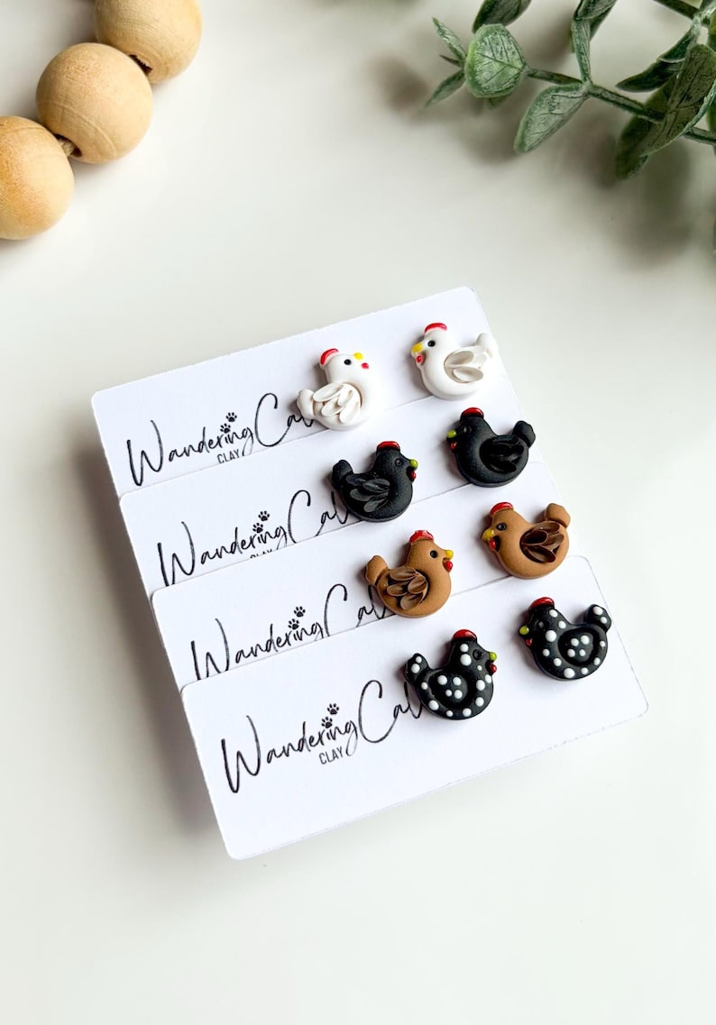 May include: A collection of handmade chicken-shaped earrings in various colors, including white, black, and brown, with red combs and yellow beaks. The earrings are displayed on white cards with the text "Wandering Clay".