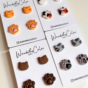 May include: A collection of cat-themed stud earrings on white cards. Designs include orange tabby cat faces, cat paws, and cat faces in various colors. The cards have the text "Wandering Calico Clay" and an Instagram handle.