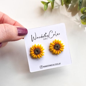 Puede incluir: Un par de pendientes de girasol hechos de arcilla polimérica. Los pendientes son amarillos con centros marrones y se muestran en una tarjeta blanca con el texto "Wandering Calico Clay" y un nombre de usuario de Instagram.
