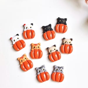 May include: A collection of cat-shaped embellishments, each nestled in a bright orange pumpkin. The cats have various colors, including black, white, gray, and orange. These decorative pieces are perfect for Halloween or fall-themed crafts.