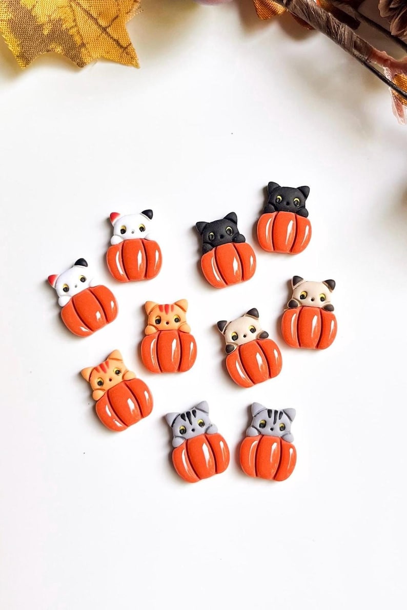 May include: A collection of cat-shaped embellishments, each nestled in a bright orange pumpkin. The cats have various colors, including black, white, gray, and orange. These decorative pieces are perfect for Halloween or fall-themed crafts.