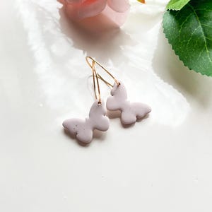May include: A pair of delicate earrings featuring a light purple butterfly charm on each earring. The earrings are made with gold-toned hoops and are perfect for adding a touch of whimsy to any outfit.
