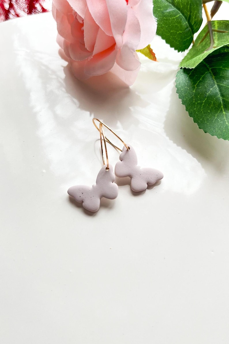 May include: A pair of delicate earrings featuring a light purple butterfly charm on each earring. The earrings are made with gold-toned hoops and are perfect for adding a touch of whimsy to any outfit.