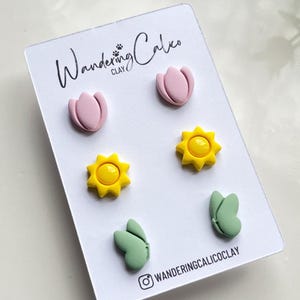 Puede incluir: Un conjunto de seis pendientes de arcilla polim&eacute;rica con una tulipa rosa, un sol amarillo y una mariposa verde. Los pendientes se muestran en una tarjeta blanca con el texto "Wandering Calico Clay".