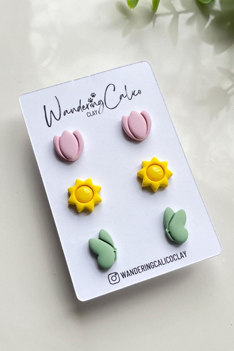 Puede incluir: Un conjunto de seis pendientes de arcilla polim&eacute;rica con una tulipa rosa, un sol amarillo y una mariposa verde. Los pendientes se muestran en una tarjeta blanca con el texto "Wandering Calico Clay".