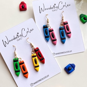 May include: A pair of earrings featuring two miniature clay crayons in green and orange, and another pair in yellow and red. The earrings are on white cards with the text "Wandering Calico Clay" and the Instagram handle "wanderingcalicoclay".