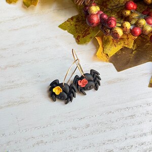 May include: A pair of black spider earrings with gold-colored hoops. Each spider has a small flower accent, one yellow and one red. The earrings are set against a white wooden background with fall foliage.