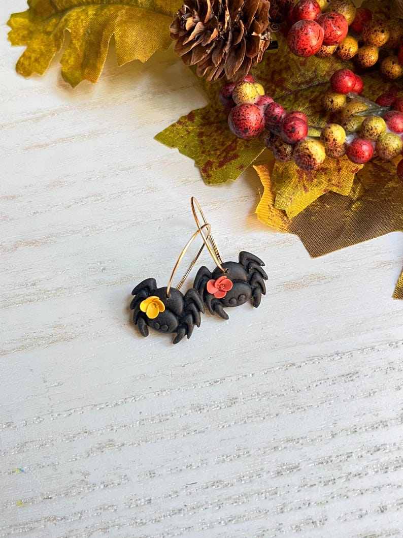May include: A pair of black spider earrings with gold-colored hoops. Each spider has a small flower accent, one yellow and one red. The earrings are set against a white wooden background with fall foliage.
