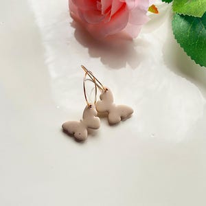 May include: A pair of gold hoop earrings with a beige butterfly charm on each hoop.