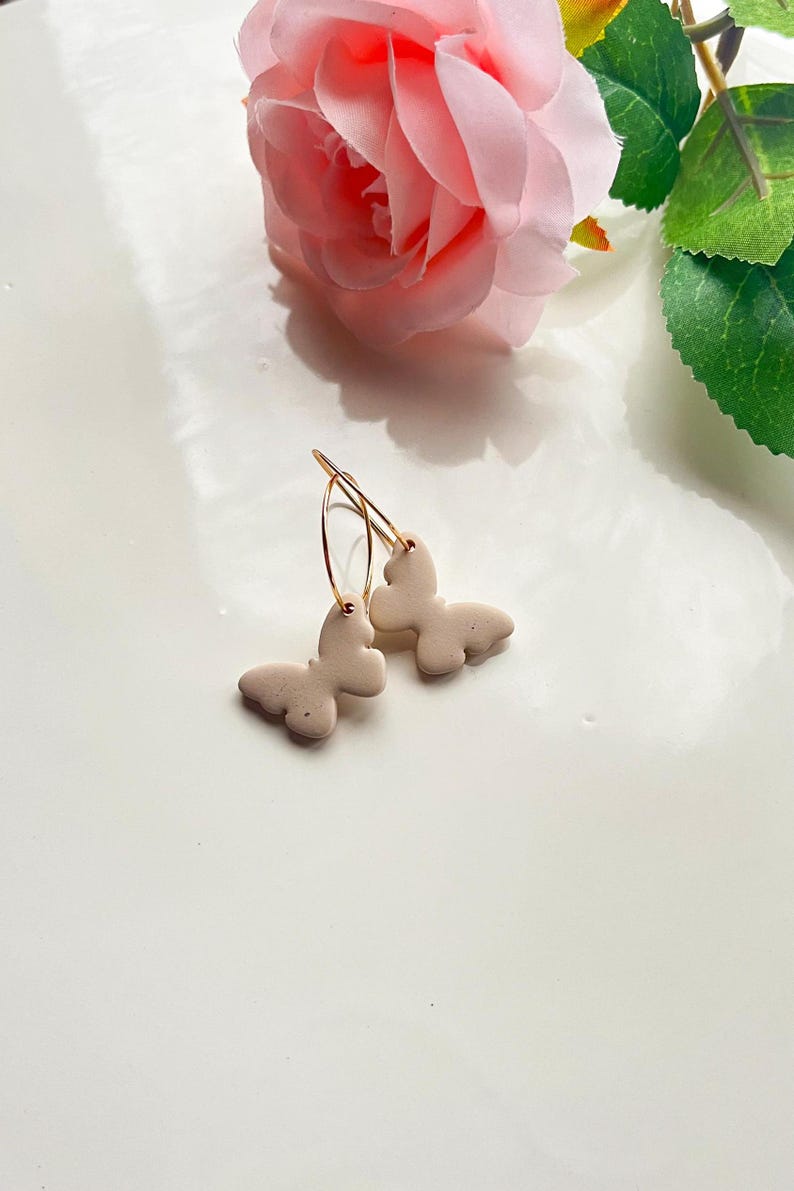 May include: A pair of gold hoop earrings with a beige butterfly charm on each hoop.