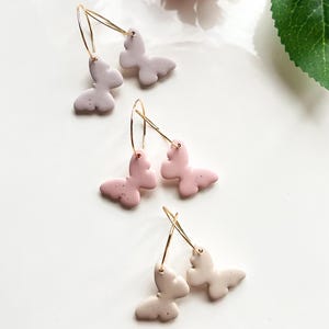 May include: Three pairs of gold hoop earrings with butterfly charms. The butterflies are made of clay and come in three colours: light pink, light purple, and white.