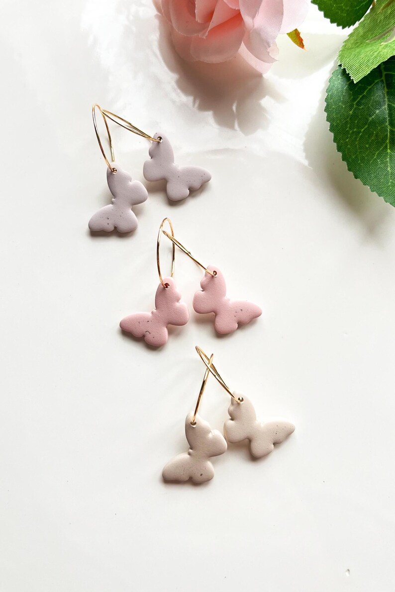 May include: Three pairs of gold hoop earrings with butterfly charms. The butterflies are made of clay and come in three colours: light pink, light purple, and white.