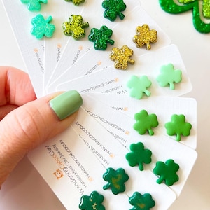 Könnte beinhalten: Ein Set mit 10 Paaren Kleeblatt-Ohrringen, handgefertigt aus Ton. Die Ohrringe sind in verschiedenen Grüntönen und mit goldenem Glitzer verziert. Die Ohrringe werden auf weißen Karten mit dem Text "WanderingCalicoClay" und "Handgefertigte Ton-Schmuckstücke" präsentiert.