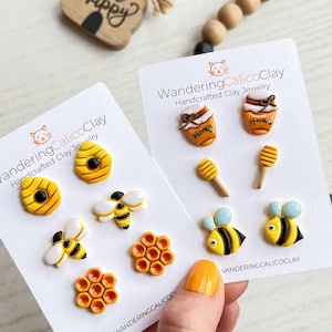 Op de afbeelding: Twee sets bijen-thema polymer clay oorbellen. De eerste set bevat een honingraat, een bij en een kleinere honingraat. De tweede set bevat een honingpot, een honinglepeltje en twee bijen. De kaarten zijn wit met zwarte tekst die "WanderingCalicoClay Handgemaakte Klei Sieraden" luidt.