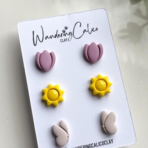 Puede incluir: Un conjunto de seis pendientes de arcilla polim&eacute;rica con una flor rosa, un sol amarillo y una mariposa gris claro. Los pendientes se muestran en una tarjeta blanca con el texto "Wandering Calico CLAY" y un icono de redes sociales.
