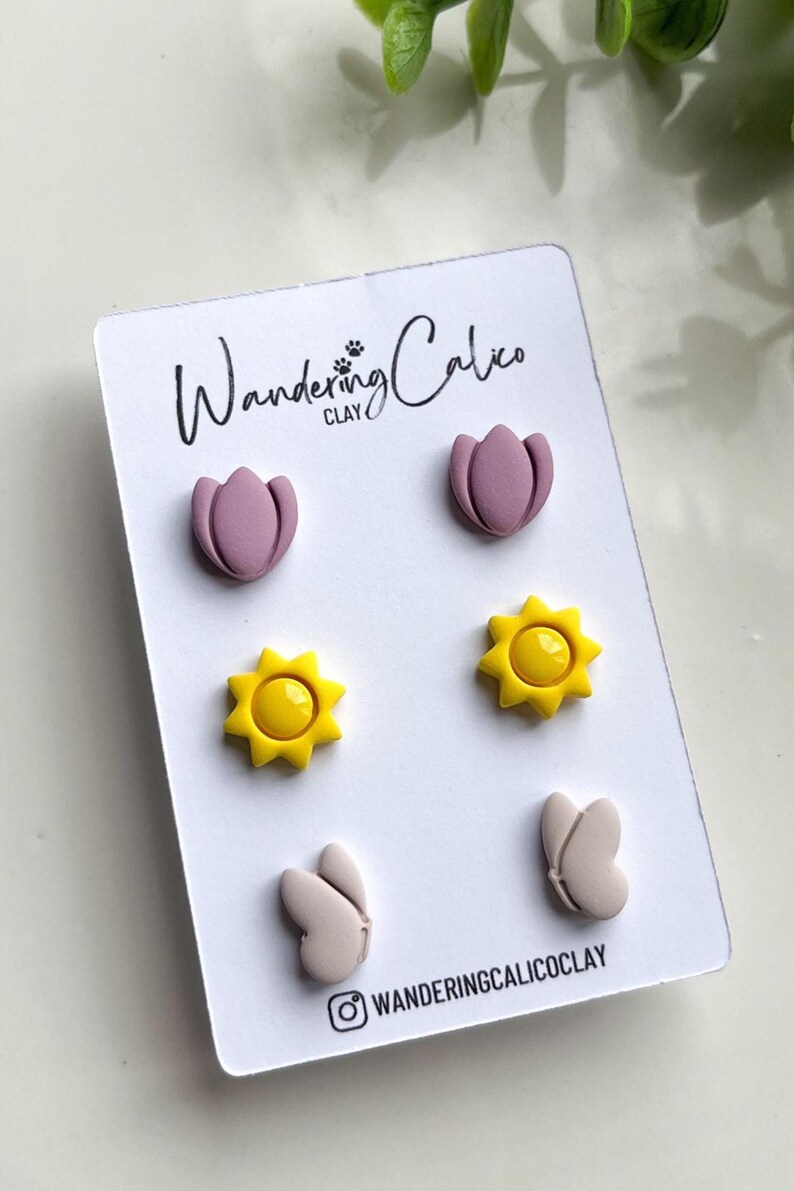 Puede incluir: Un conjunto de seis pendientes de arcilla polim&eacute;rica con una flor rosa, un sol amarillo y una mariposa gris claro. Los pendientes se muestran en una tarjeta blanca con el texto "Wandering Calico CLAY" y un icono de redes sociales.