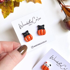May include: Handmade Halloween-themed earrings featuring black cat figures sitting in orange pumpkins. The earrings are displayed on white cards with the text "Wandering Calico Clay" and the Instagram handle. Fall-themed decor surrounds the earrings.