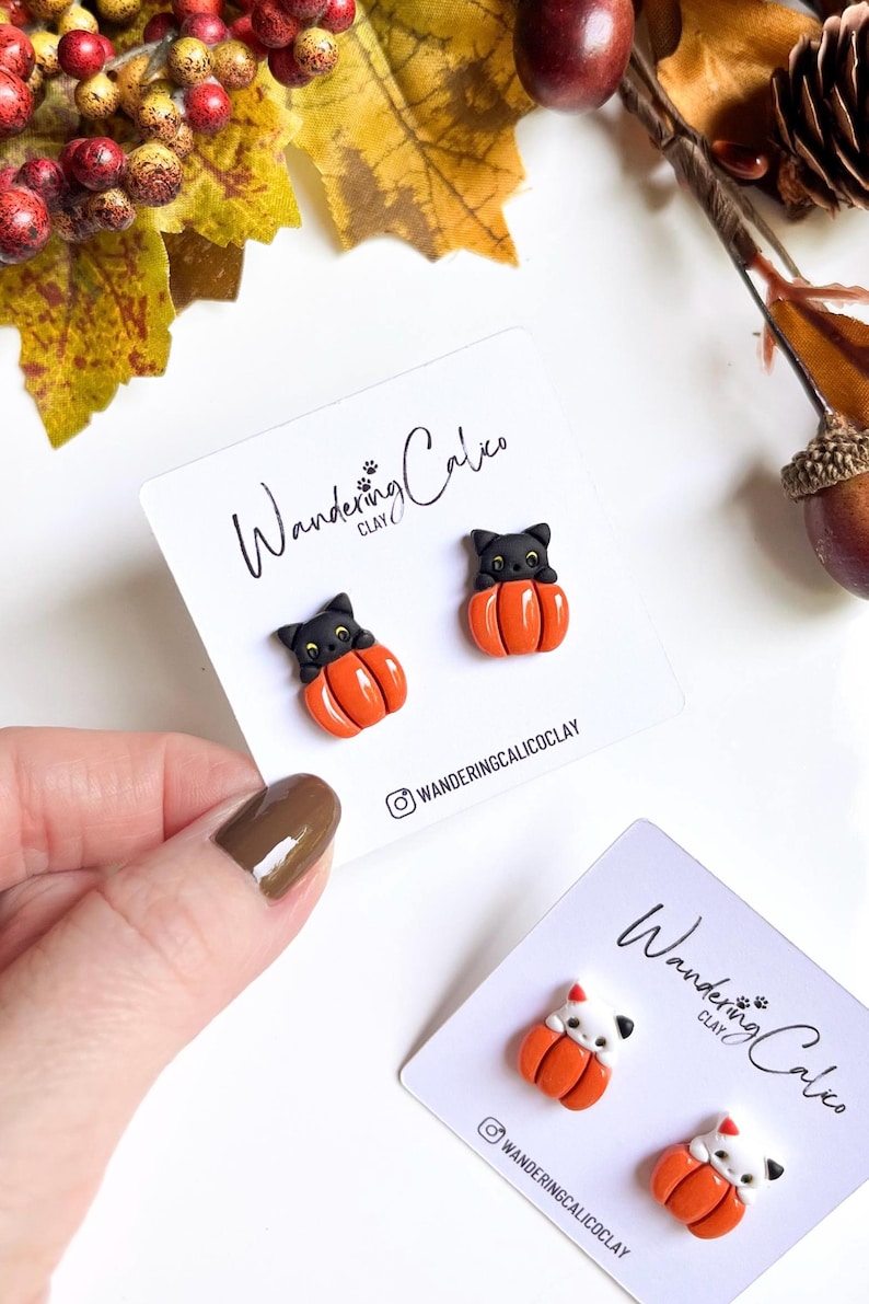 May include: Handmade Halloween-themed earrings featuring black cat figures sitting in orange pumpkins. The earrings are displayed on white cards with the text "Wandering Calico Clay" and the Instagram handle. Fall-themed decor surrounds the earrings.