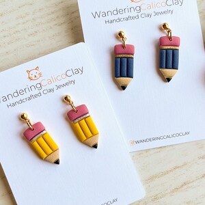 May include: Handcrafted clay jewelry featuring pencil-shaped earrings. The earrings are yellow, blue, pink, and black, with gold-colored accents. The earrings are displayed on white cards with the text "WanderingCalicoClay Handcrafted Clay Jewelry".