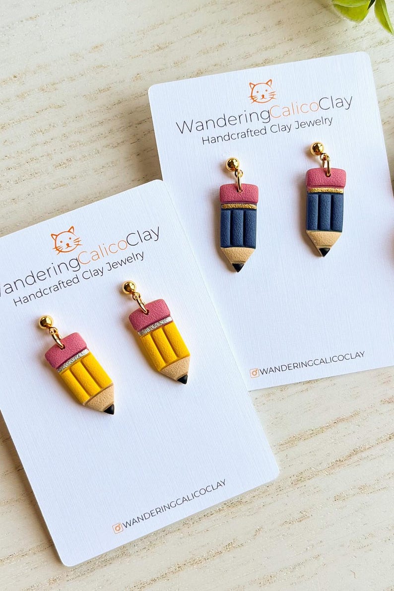 May include: Handcrafted clay jewelry featuring pencil-shaped earrings. The earrings are yellow, blue, pink, and black, with gold-colored accents. The earrings are displayed on white cards with the text "WanderingCalicoClay Handcrafted Clay Jewelry".