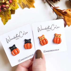 May include: Two pairs of handmade clay earrings featuring a black cat and a tabby cat inside orange pumpkins. The earrings are displayed on white cards with the text "Wandering Calico Clay" and social media handles. Autumn-themed jewelry.